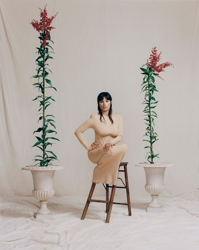 A woman in a tan dress sits on a wooden stool in a bright photo studio, flanked by tall flowers in ornate planters against a white backdrop.