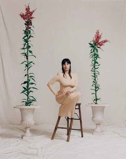 A woman in a tan dress sits on a wooden stool in a bright photo studio, flanked by tall flowers in ornate planters against a white backdrop.