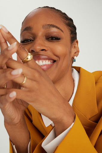Close-up of a smiling Black woman in a yellow blazer with gold rings on her fingers, posing indoors against a white background.