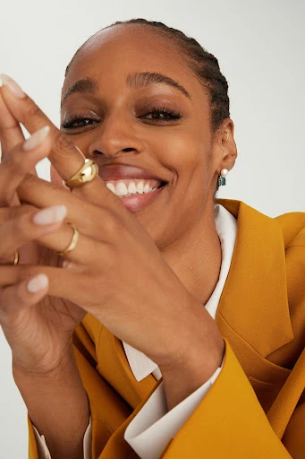 Close-up of a smiling Black woman in a yellow blazer with gold rings on her fingers, posing indoors against a white background.