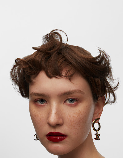 Close-up shot of a young fair-skinned adult woman with auburn hair, freckles, and red lipstick wearing gold hoop earrings against a white background.