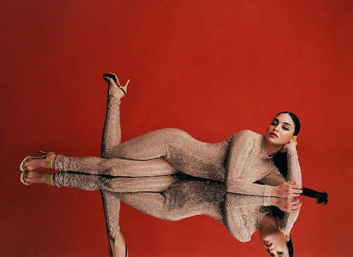 Woman in sparkly bodysuit and high heels poses on a mirrored surface against a red background in a fashion-style photograph.