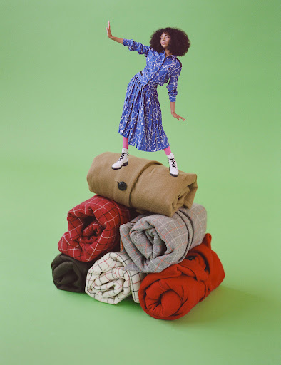 Woman in patterned dress strikes a pose on a stack of rolled blankets against a solid green background in a studio shot.