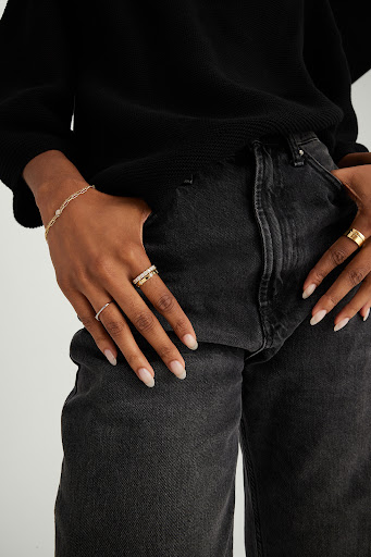 Close-up of woman's hands with multiple rings and bracelet, posing against a dark grey denim jeans and a black sweater.