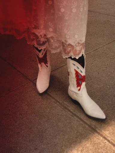 Close-up of white cowboy boots adorned with red and blue butterfly designs, paired with a white lace dress, standing on a grey surface.