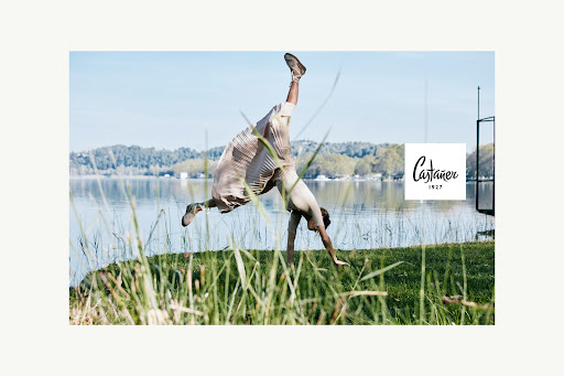 Adult female does a handstand outdoors on a grassy riverbank with trees, water, and blue sky in the background. Fashion editorial.