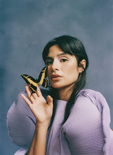 Woman with a yellow and black butterfly on her face in a portrait setting, wearing a pleated purple top against a mottled gray backdrop.