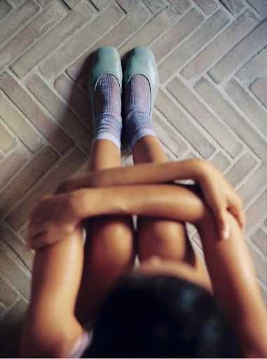 Young person wearing blue ballet shoes with sheer socks sitting with legs crossed on herringbone tile indoors, embracing knees.