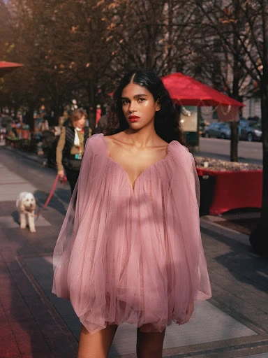 A stylish Indian woman in a pink tulle dress poses on a city street with trees, buildings, and people in the background.