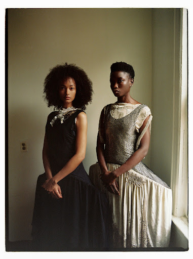 Two women pose indoors wearing dresses with vintage fashion aesthetic, in a close-up portrait with a neutral backdrop and natural light.