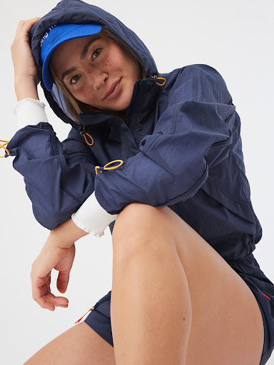 Attractive woman with fair skin and freckles poses wearing a blue hooded jacket and cap against a white background in a studio shot.
