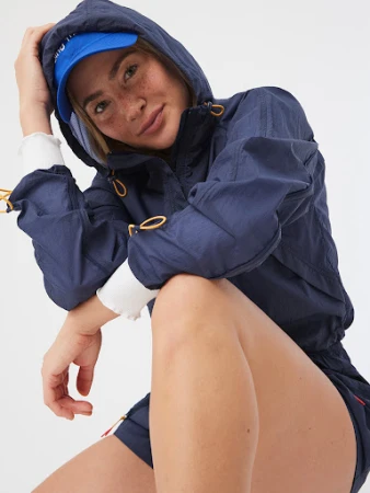 Attractive woman with fair skin and freckles poses wearing a blue hooded jacket and cap against a white background in a studio shot.
