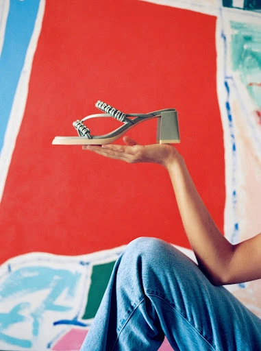 Elevated sandal is held by a hand with red abstract background and blue jeans. The sandal is light gray with a block heel.