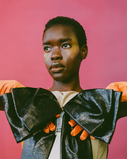 Close-up shot of an African female model in a shiny black bow-shaped top and orange gloves posing against a pink background in a photo studio.