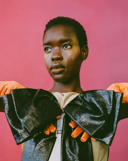 Close-up shot of an African female model in a shiny black bow-shaped top and orange gloves posing against a pink background in a photo studio.