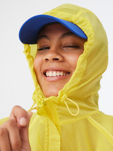 Smiling woman with natural skin and teeth wearing a yellow mesh jacket and blue baseball cap against a neutral white background.