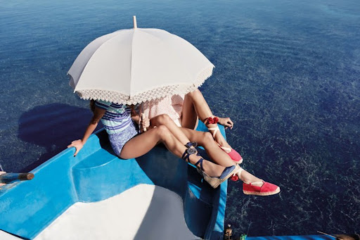 Two women sit on a blue boat, under a white parasol, wearing espadrilles, in the sunny marine environment with clear blue water.
