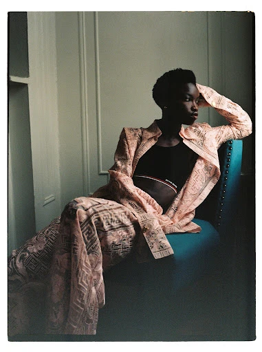Elegant Black woman in patterned blazer and pants sitting on turquoise chair in bright interior, glancing away in fashion pose.