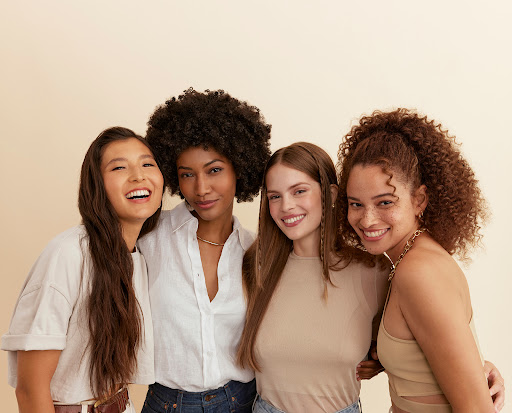 Group portrait of four diverse adult women smiling and posing together indoors in neutral casual clothing against a beige backdrop.