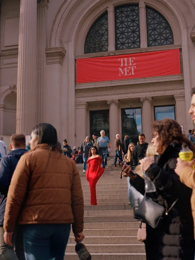 Woman in a red dress walks down the stairs of The Metropolitan Museum of Art in New York City, surrounded by a crowd of people.