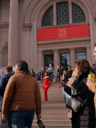 Woman in a red dress walks down the stairs of The Metropolitan Museum of Art in New York City, surrounded by a crowd of people.