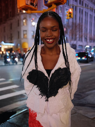 Woman with braided hair smiles, wearing a black and white jacket, stands on a city street with traffic lights and buildings in the background.