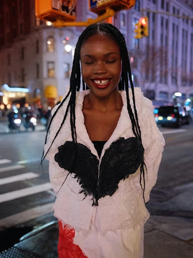 Woman with braided hair smiles, wearing a black and white jacket, stands on a city street with traffic lights and buildings in the background.