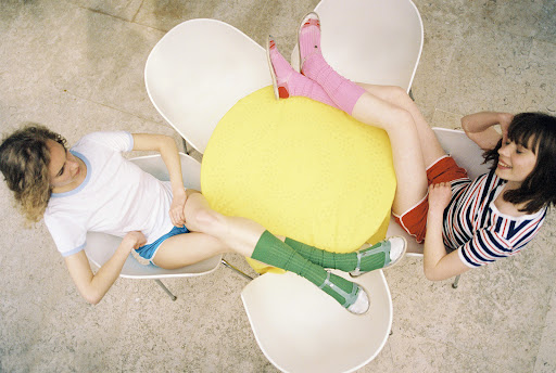 Two young women sit on chairs around a yellow lemon-shaped cushion, wearing colorful shorts and socks in an overhead shot.