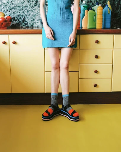 Unidentifiable woman stands in a blue dress, patterned socks, and platform sandals in front of yellow cabinets and kitchenware.
