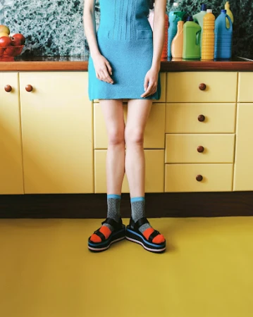 Unidentifiable woman stands in a blue dress, patterned socks, and platform sandals in front of yellow cabinets and kitchenware.