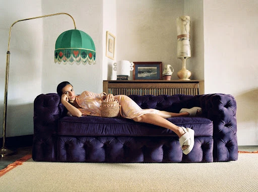 Woman in peach dress reclines on a purple tufted couch in an indoor setting with a green fringed lamp and woven handbag.