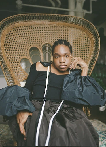 Portrait of a woman in a large wicker chair wearing a black top and brown skirt with white decorative straps, indoor environment.