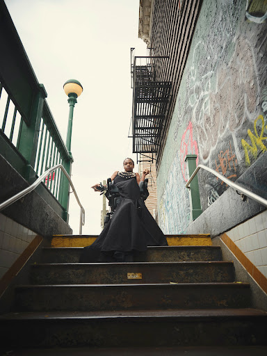 Adult female in a dramatic black dress poses on subway stairs in an urban setting with graffiti-covered walls and a vintage street lamp.