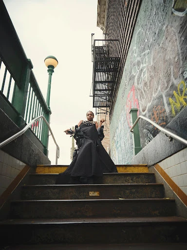 Adult female in a dramatic black dress poses on subway stairs in an urban setting with graffiti-covered walls and a vintage street lamp.