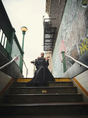 Adult female in a dramatic black dress poses on subway stairs in an urban setting with graffiti-covered walls and a vintage street lamp.