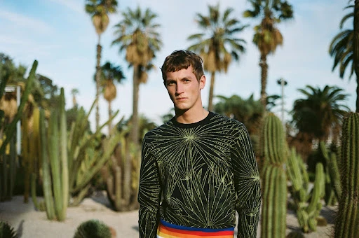 A light-skinned adult male standing in a desert botanical garden surrounded by tall cacti and palm trees with a clear blue sky.