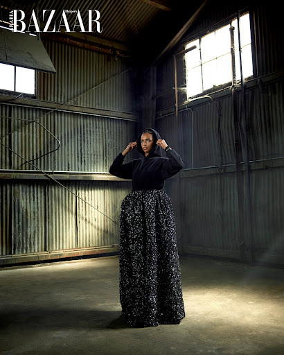 Fashion editorial shot of a Black woman in a hooded sweatshirt and long skirt, posing in an industrial setting with natural lighting.