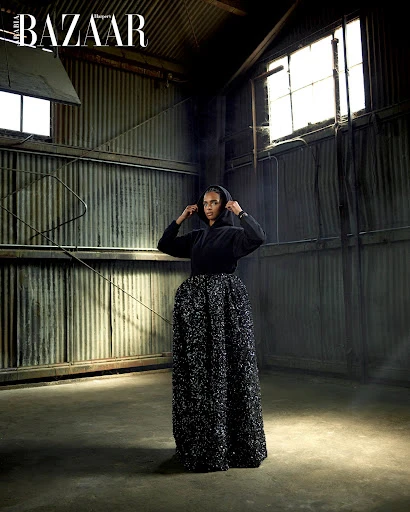 Fashion editorial shot of a Black woman in a hooded sweatshirt and long skirt, posing in an industrial setting with natural lighting.