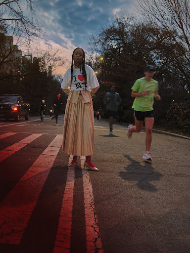 Woman in pleated skirt with 'I Love NY' shirt poses on crosswalk in city park at twilight. Joggers in the background.