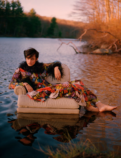 Person in floral dress and feathered collar lounges on sofa in lake at sunset, surrounded by forest in an outdoor editorial fashion shot.