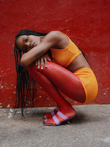 Black woman with long braids wearing an orange crop top and shorts while squatting against a textured red wall