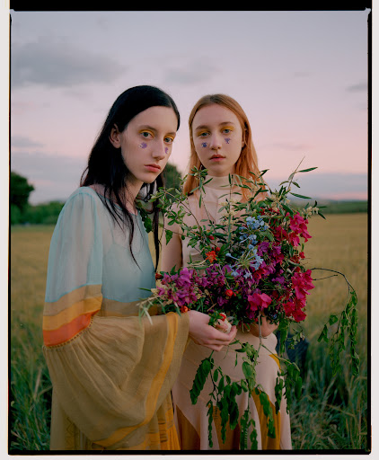 Two young women stand in a field of wheat, holding colorful wildflowers, wearing dresses, and heart shaped face decoration, against a dusk sky.