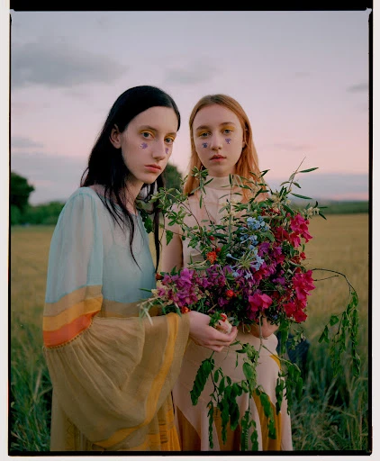 Two young women stand in a field of wheat, holding colorful wildflowers, wearing dresses, and heart shaped face decoration, against a dusk sky.
