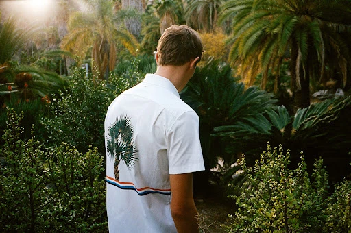 Back view of an adult man in a white patterned shirt standing amid lush green tropical foliage and palm trees on a bright day.