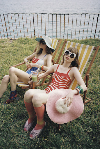 Two women in swimsuits relax on deck chairs in a grassy outdoor setting with the ocean in the background, wearing hats and sunglasses.