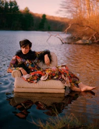 Model wearing a floral patterned dress with dark feather trim, seated on a cushioned armchair placed in a calm lake.