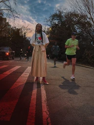 Woman in an I Love NY graphic t-shirt and pleated tan midi skirt standing on an asphalt street at dusk.