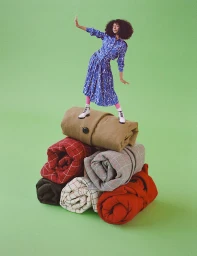 Model with afro hair in blue patterned outfit posing atop a precarious stack of rolled garments against green backdrop