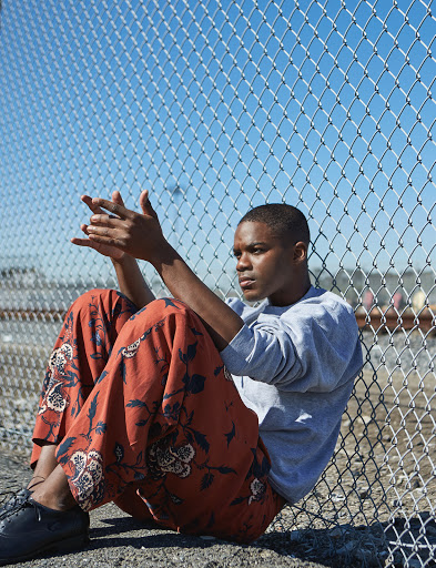 Young Black man with short hair sits near a chain link fence wearing a grey sweatshirt and floral print pants in an outdoor setting.