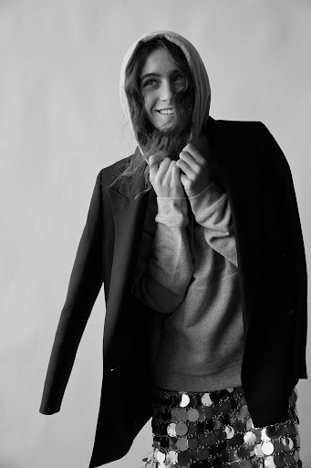 Black and white portrait of a smiling adult woman in a hoodie, jacket, and sequin skirt, posing against a neutral studio backdrop.